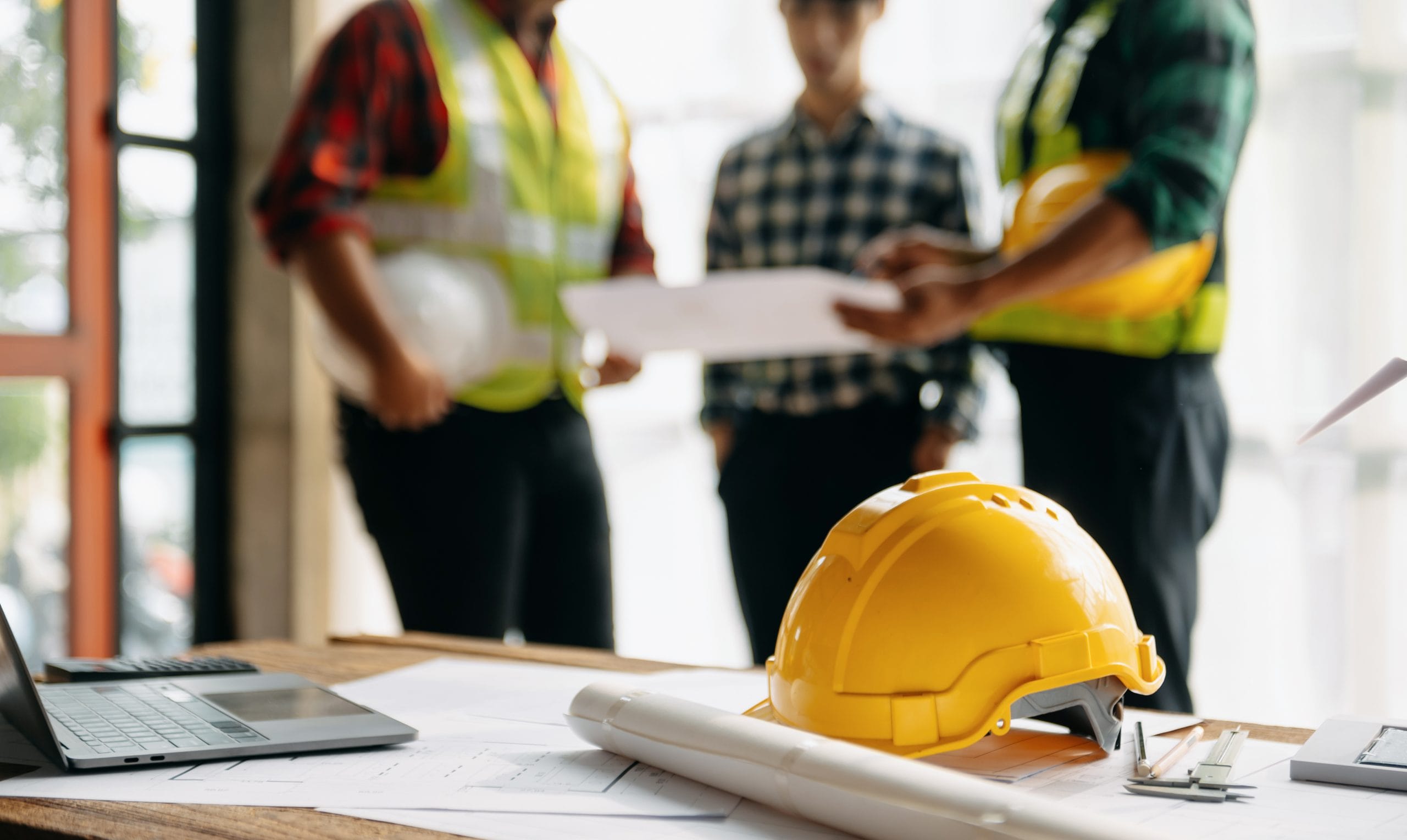 Construction site supervisors reviewing drawings in the background, with a hard hat and laptop on the table in the foreground.